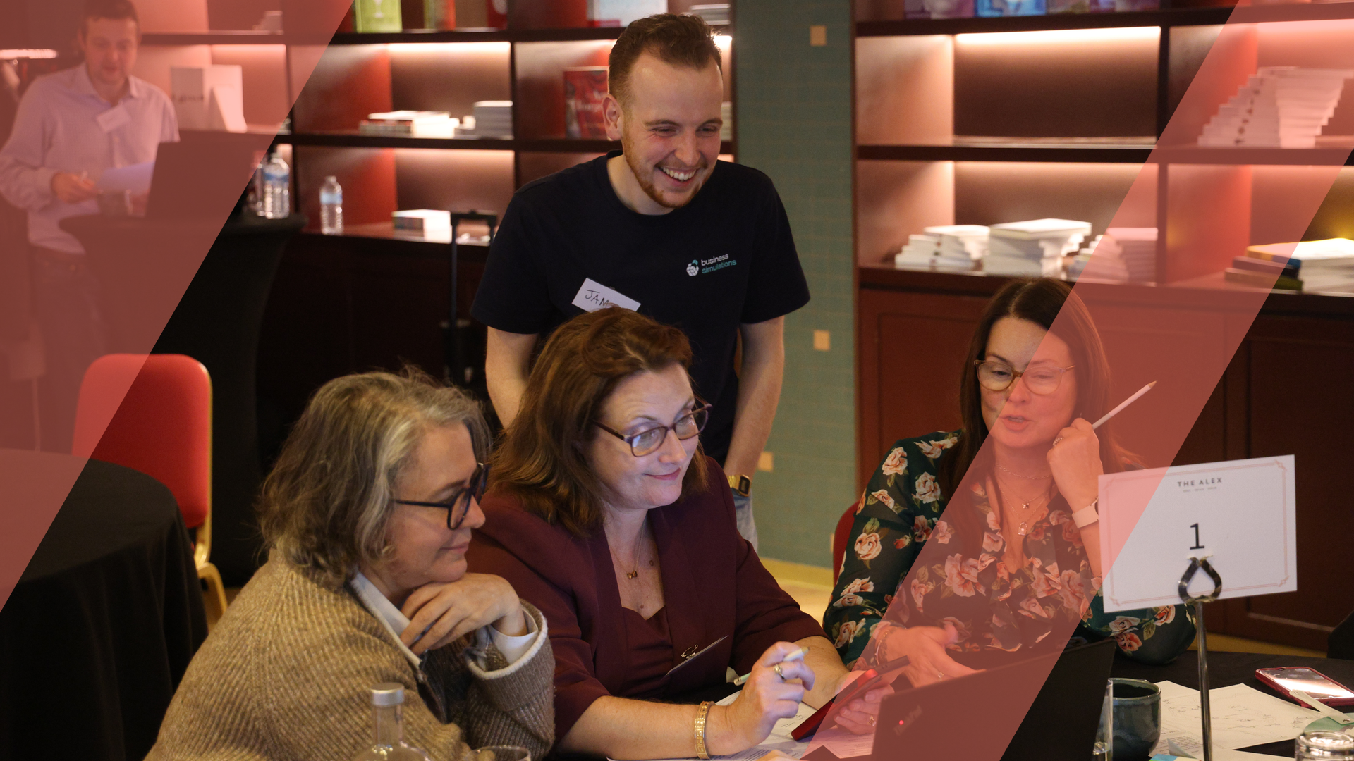 A colleague leaning in to help two teammates working at a laptop during a live business simulation, overlaid with an orange Business Simulations brand motif