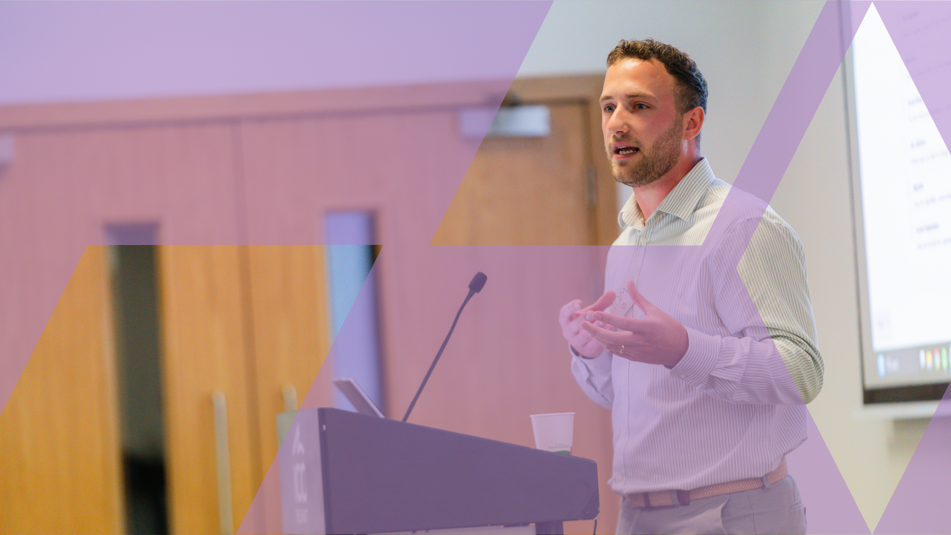 A facilitator leading a live, in-person management development session, gesturing as he speaks beside a lectern, overlaid with a purple Business Simulations brand motif