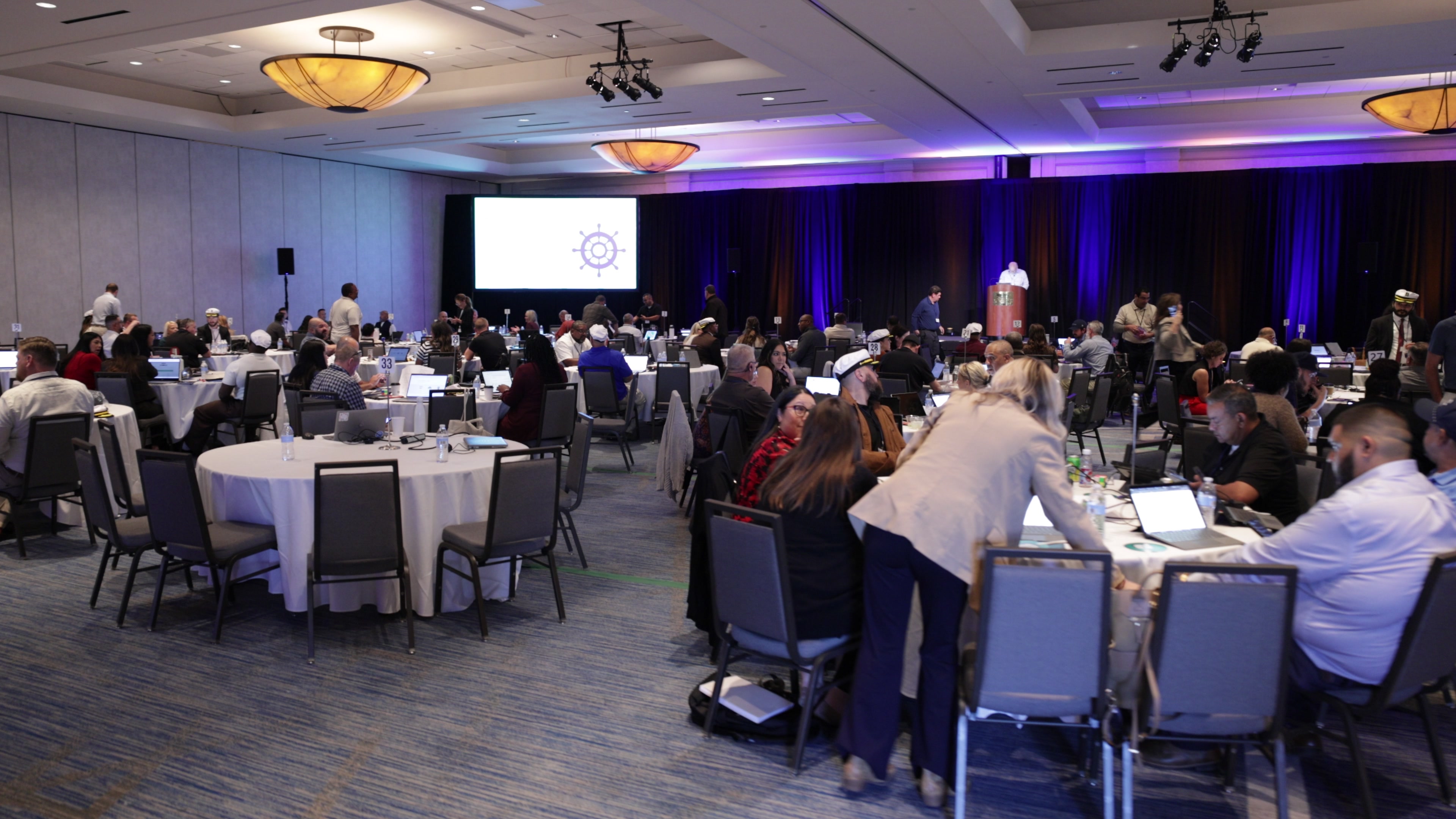 A wide view of a large conference ballroom where many people are seated at round tables using laptops, with a person at a podium in the background and a large screen showing a ship's wheel logo.