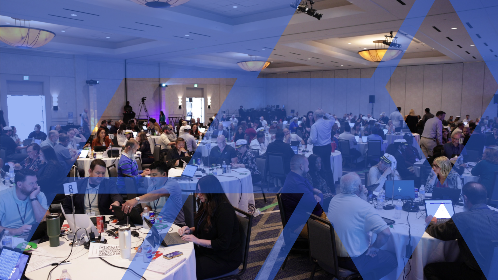 A wide view of a large conference room where teams work at round tables, one participant pointing at a shared screen.