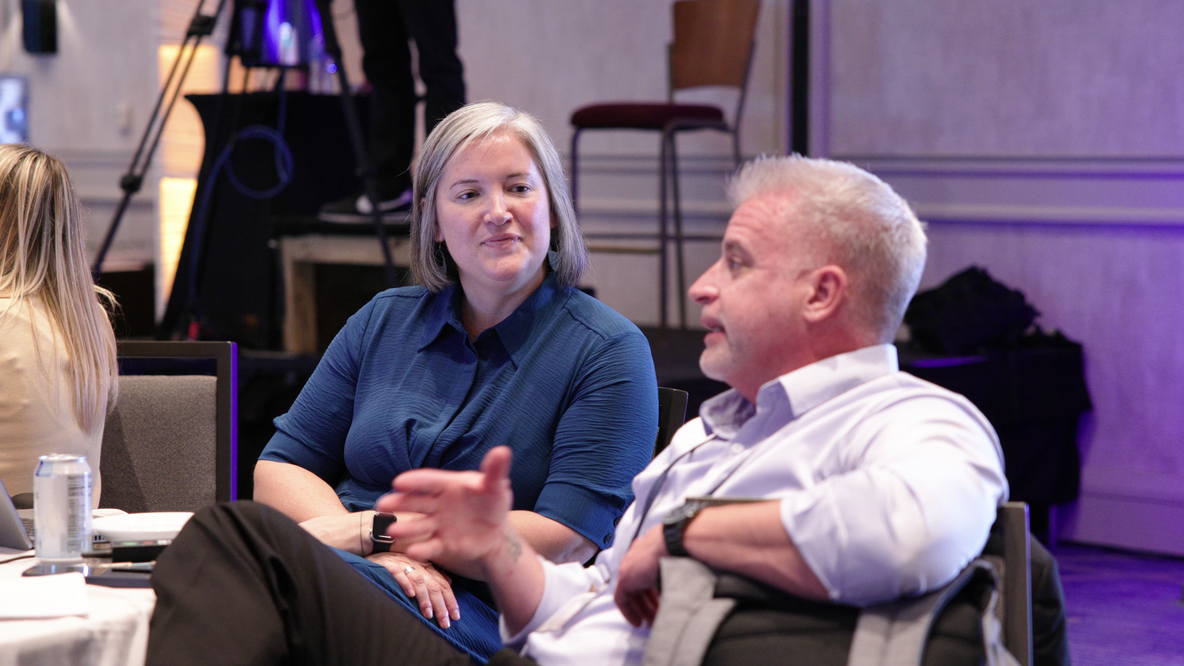 A woman and a man sit at a round table in a conference room, looking attentively to the right.